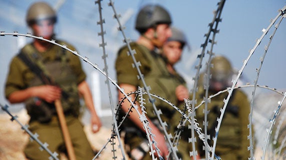 Armed soldiers standing guard in the distance, with the camera focused on barbed-wire in the foreground.